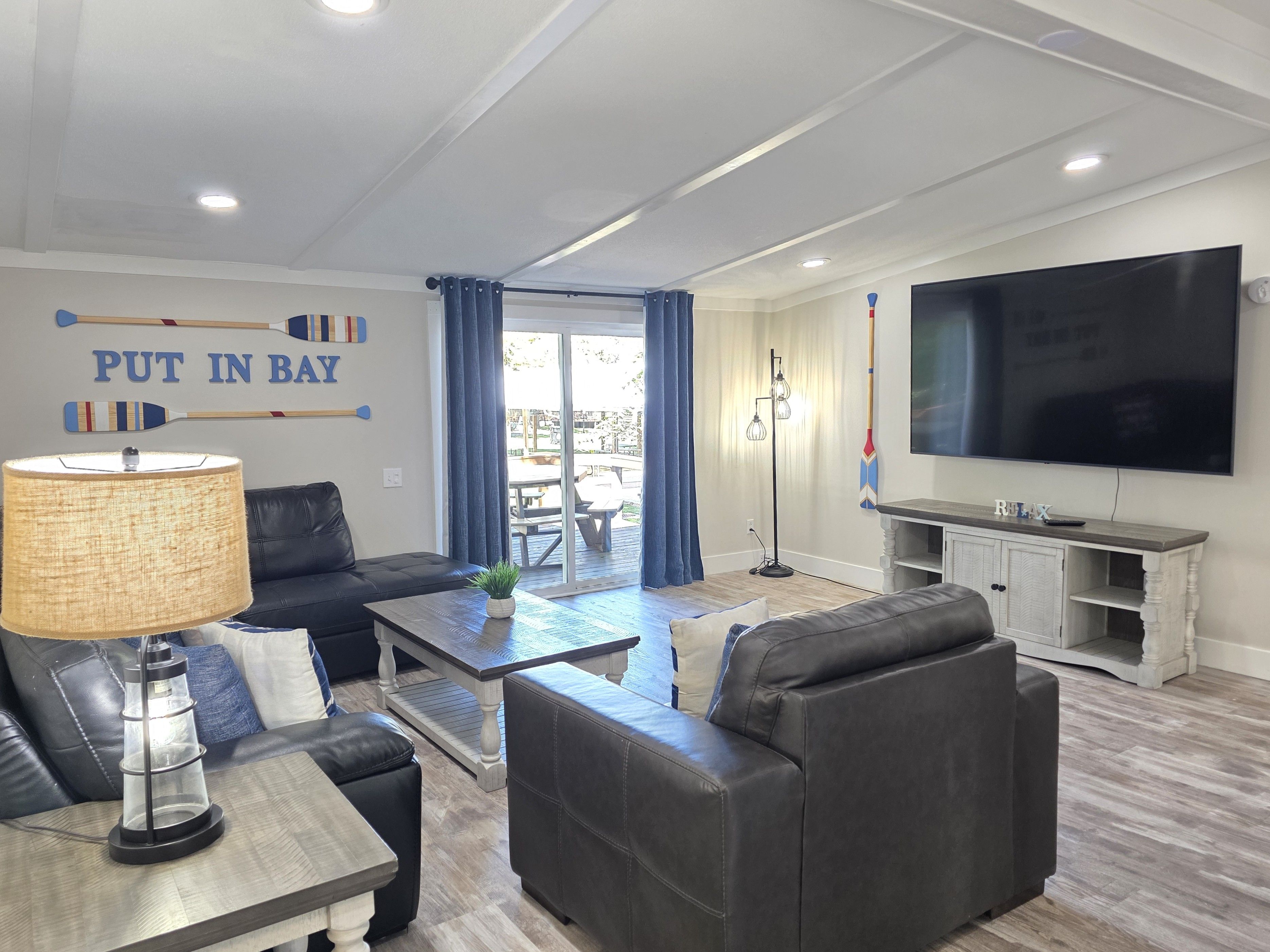 A modern living room with a sofa, coffee table, and a TV. There's a wall with a 'PUT IN BAY' sign and a large window with curtains.