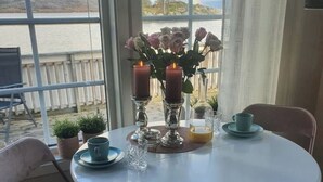 Et rundt bord med to lys, en vase med blomster og et glas appelsinjuice. Der er to stole ved bordet, og et vindue med udsigt over vand og bakker.