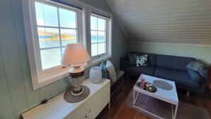 Et hyggeligt rum med et stort vindue, en lampe på et hvidt skab, en mørkeblå sofa og et lille hvidt bord.