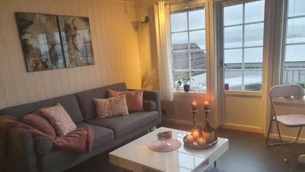 En moderne stue med en grå sofa, et hvidt sofabord med lys og udsigt til byen gennem store vinduer.