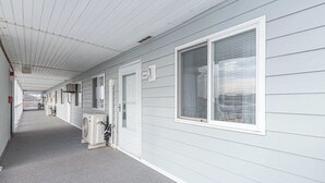 Un pasillo con una cubierta, unidades de aire acondicionado y una fila de habitaciones con ventanas y puertas.
