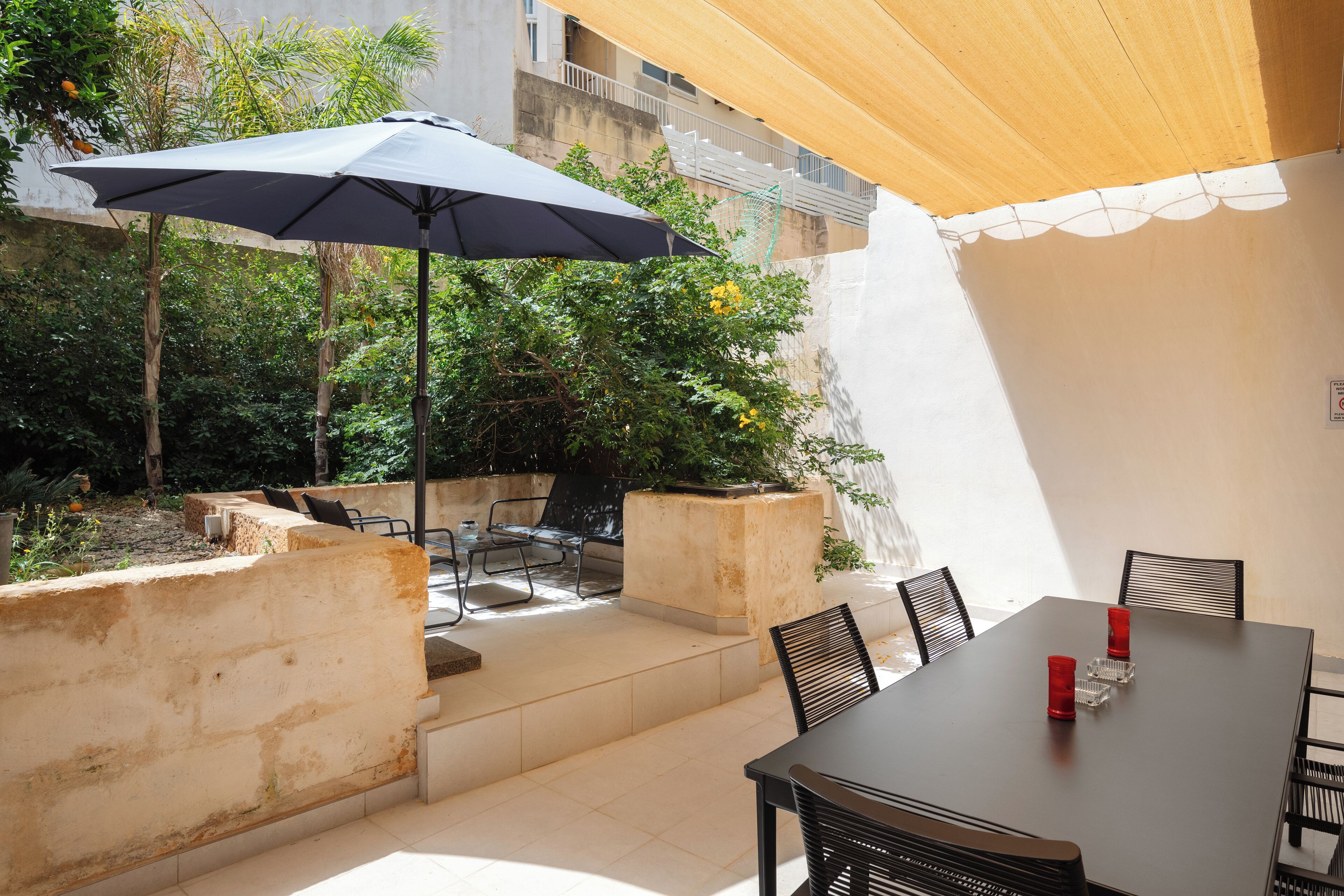 Un patio exterior con una mesa y sillas, una sombrilla grande y vistas a un jardín con plantas y un muro.