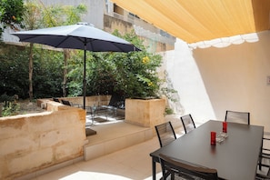 Um pátio ao ar livre com uma mesa e cadeiras, um grande guarda-sol e vista para um jardim com plantas e um muro.