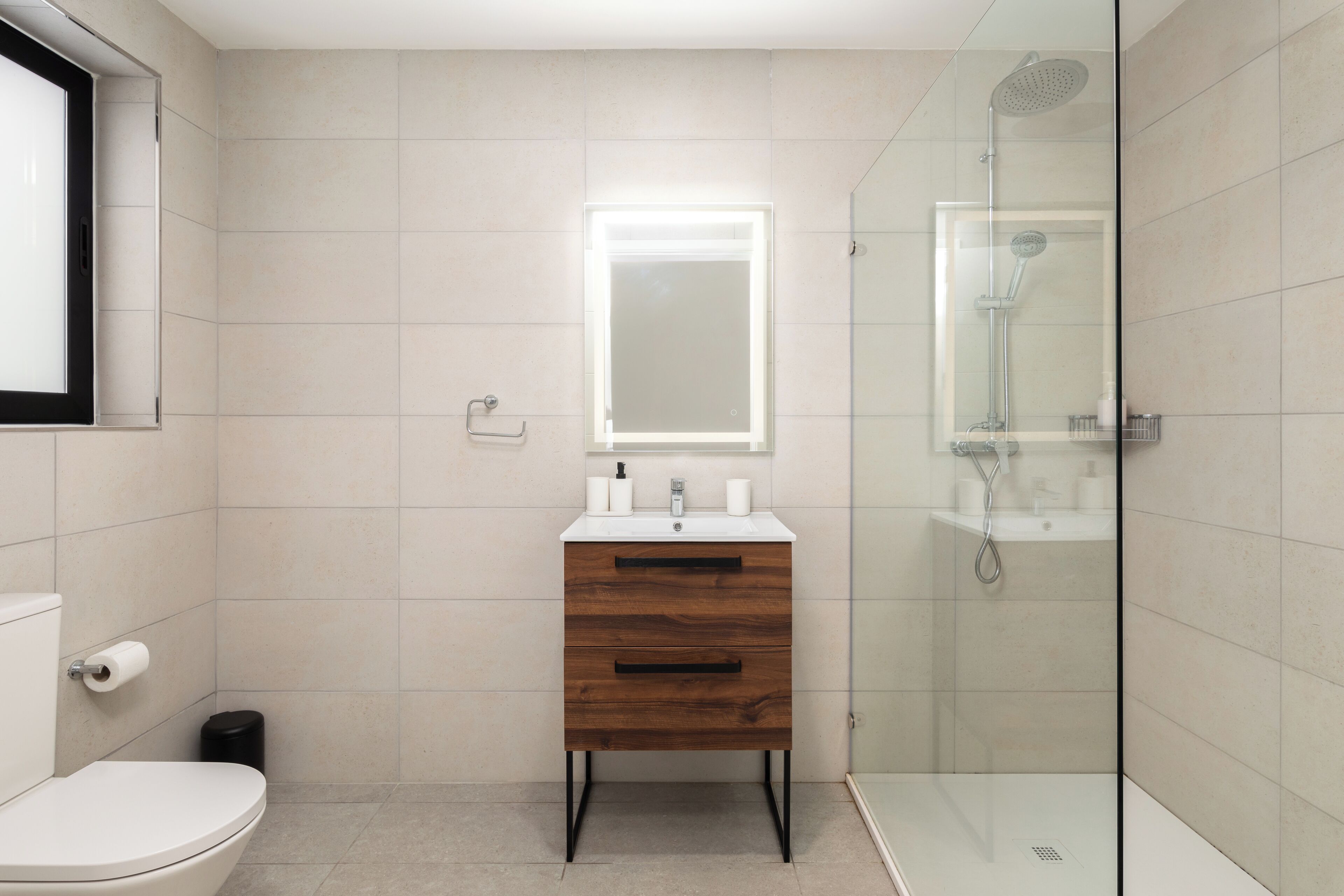 Un baño moderno con inodoro, un mueble de tocador de madera con espejo y una zona de ducha con mampara de vidrio.