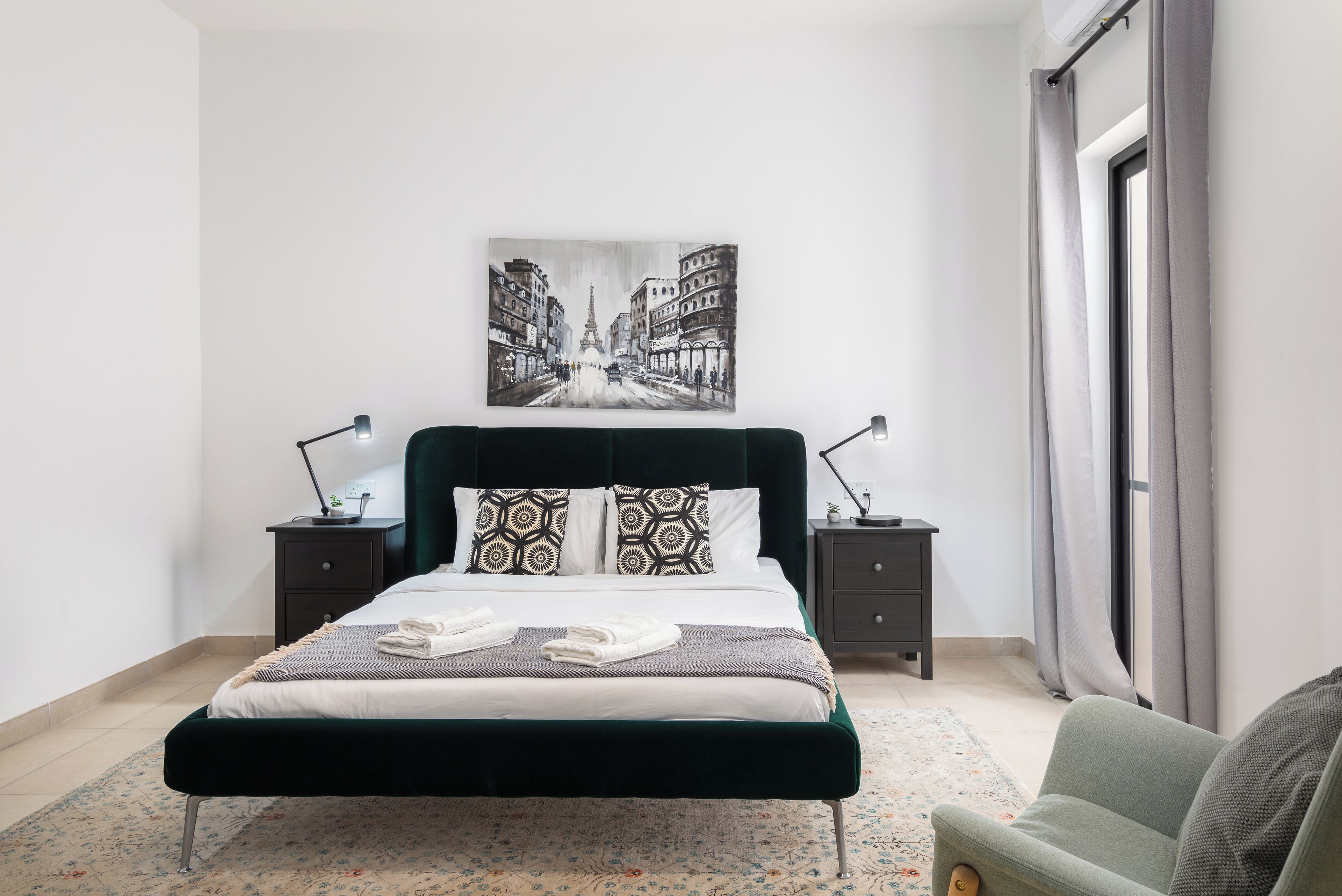 A neatly made bed with a cityscape painting above it, a bedside table with a lamp, and a view of a city street through the window.