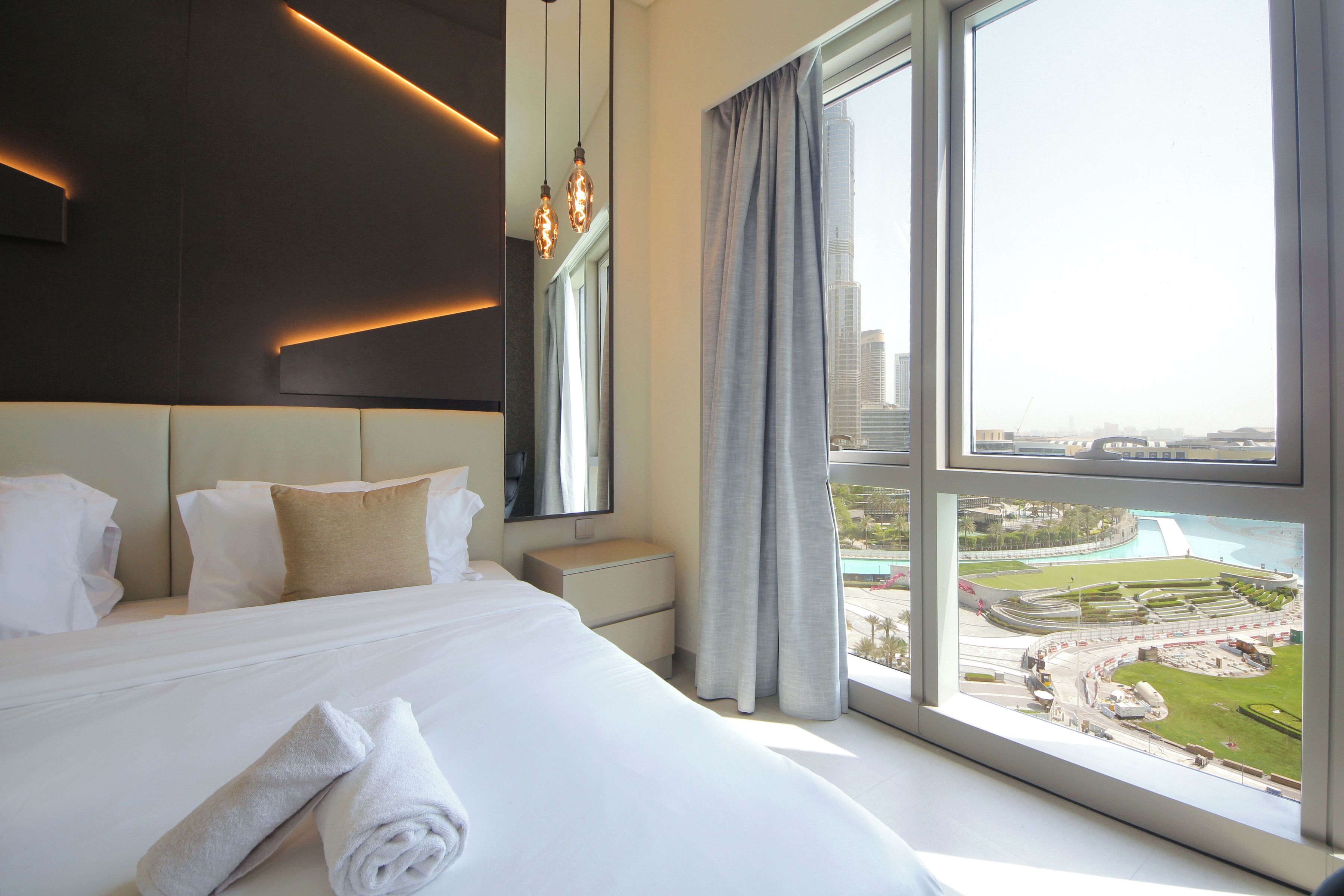 A modern hotel room with a large bed, a nightstand, a mirror, and a view of a cityscape through large windows.