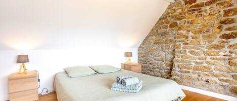 A bedroom with a stone wall, a wooden floor, a bed with a white bedspread, two bedside tables with lamps, and a folded towel on the bed.