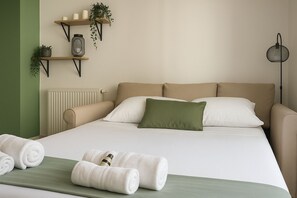 A neatly made bed with white linens and a green pillow, a beige sofa, and a wall-mounted shelf with plants and candles. - Charming apartment - Near train station and town center (MontPellier)