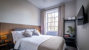 A neatly arranged bedroom with a bed, bedside table, lamp, and a shelf with a plant and books. - Dalton Sq Apartments (Lancaster)