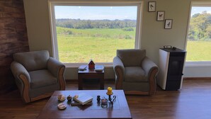 Una habitación con dos sillones, una mesita con adornos y vistas a un campo verde a través de grandes ventanales.