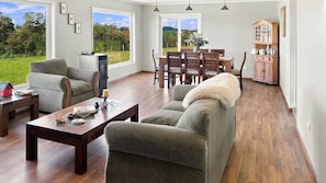 A spacious living room with a sofa, a dining table, and chairs, large windows with a view of the outdoors, and a wooden cabinet. - Oceana Suites Sueño Rural (Puerto Varas)