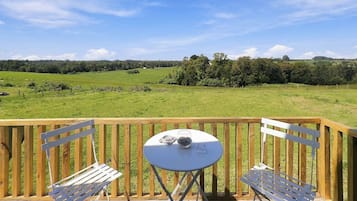 A wooden deck with a table and two chairs, overlooking a green field and trees.