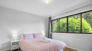 Un dormitorio con un ventanal grande, una cama con ropa de cama a cuadros, dos mesitas de noche con lámparas y vistas al exterior con vegetación.