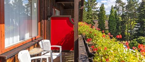 A wooden deck with two white chairs and a table, overlooking a garden with red flowers and tall trees.
