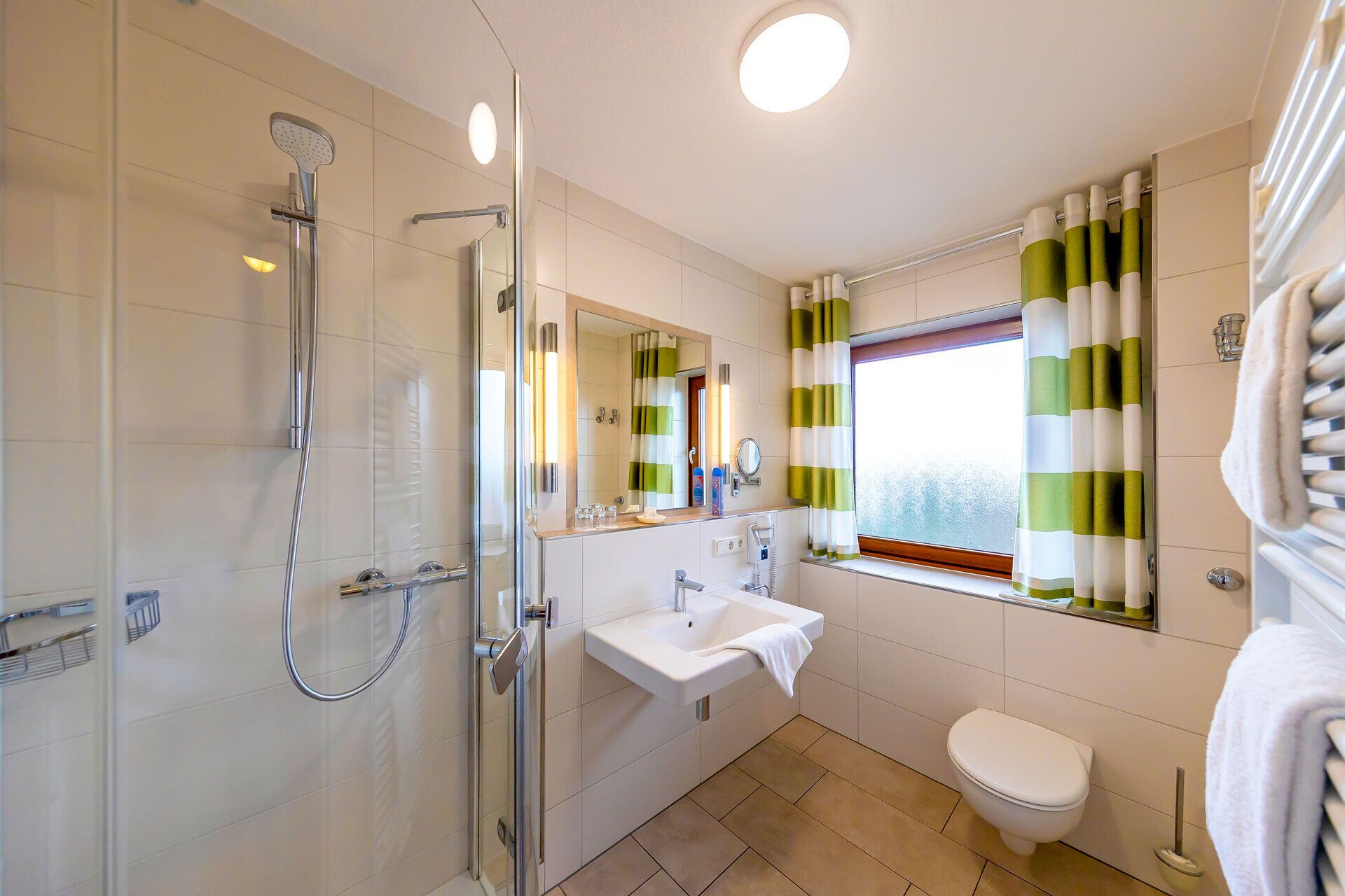 Een moderne badkamer met een douchecabine van glas, een wastafel, een toilet en een raam met gestreepte gordijnen.