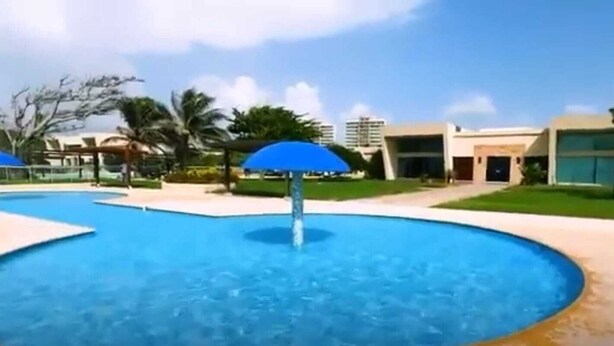 A swimming pool with a blue umbrella, surrounded by palm trees and modern buildings. - RESORT FRENTE AL MAR (Juan de Acosta)