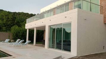 Un edificio moderno con fachada de vidrio, un área de piscina con sillones reclinables y una terraza de madera.