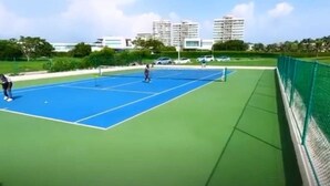 Una cancha de tenis al aire libre con una valla verde y edificios al fondo.