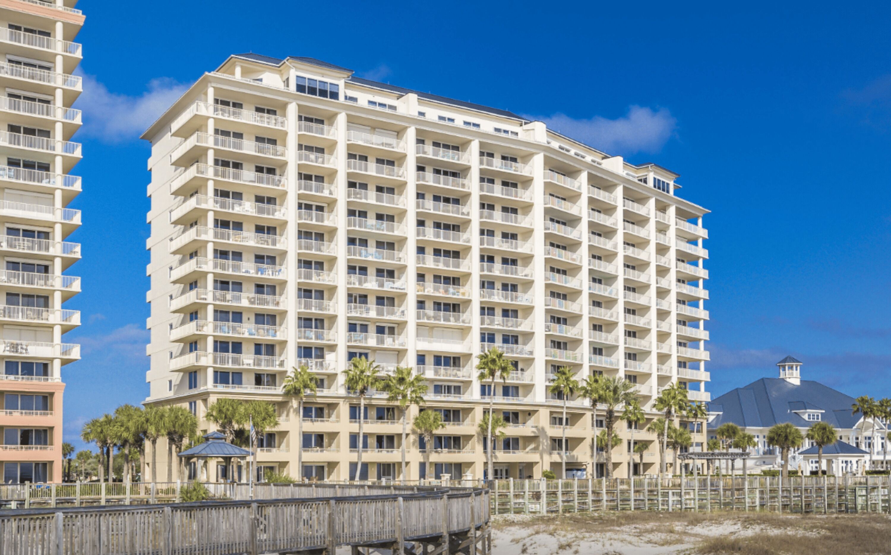Un hotel de varios pisos con balcones, situado cerca de una playa con palmeras y un paseo marítimo de madera.