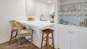 Eine moderne Küche mit weißer Arbeitsplatte, zwei Barhocker, einer Spüle und einem Glastrennregal mit Dekoration.