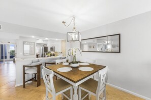 A modern dining area with a wooden table, white chairs, and a central vase with flowers. - The Somerset Solace | Pools, Tennis Courts & Gym (Marco Island)