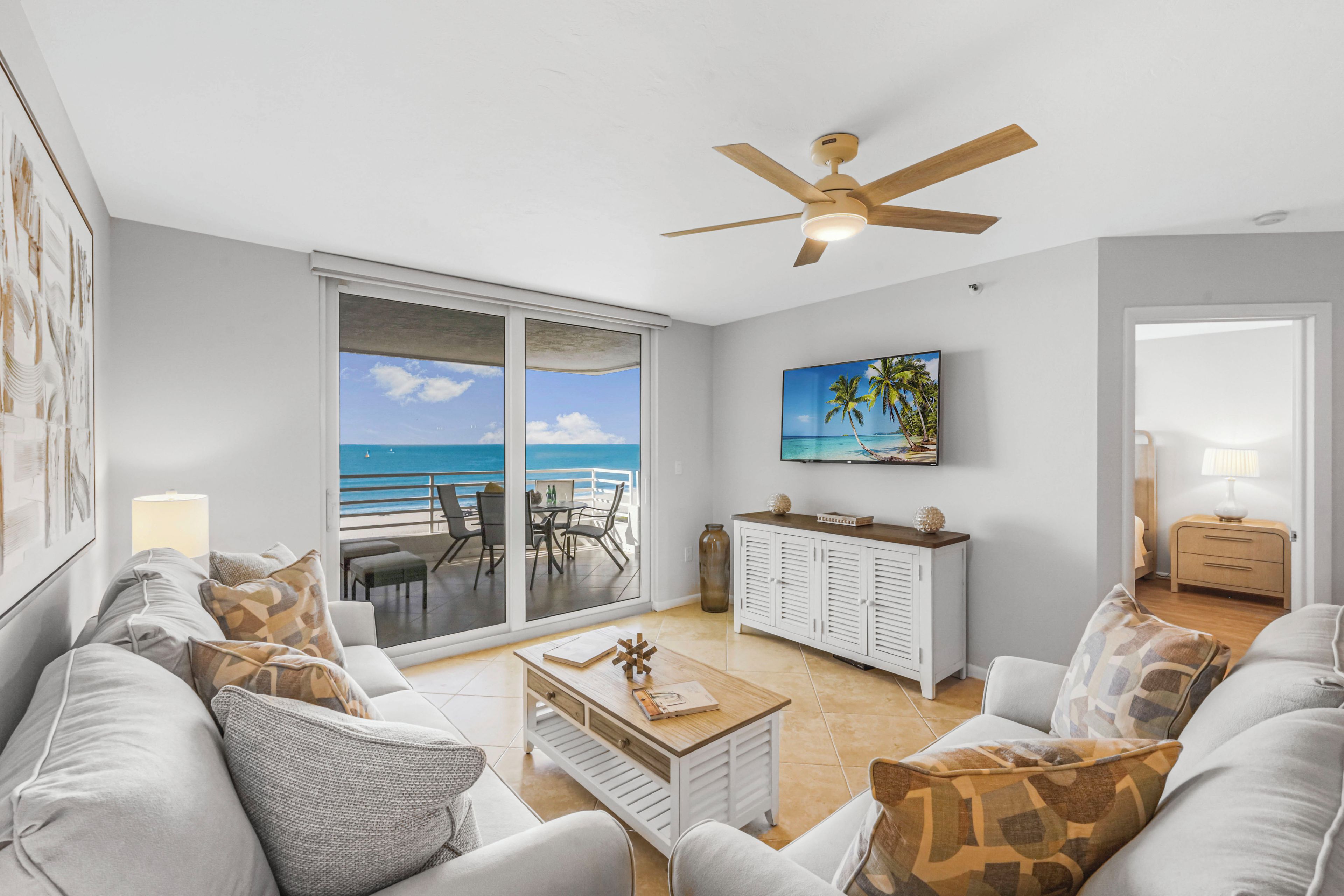 A living room with a view of the ocean, a ceiling fan, a coffee table, and a TV on the wall.