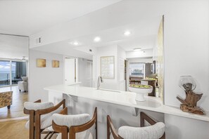 A modern kitchen with a white countertop, bar stools, and a large mirror. - The Somerset Solace | Pools, Tennis Courts & Gym (Marco Island)