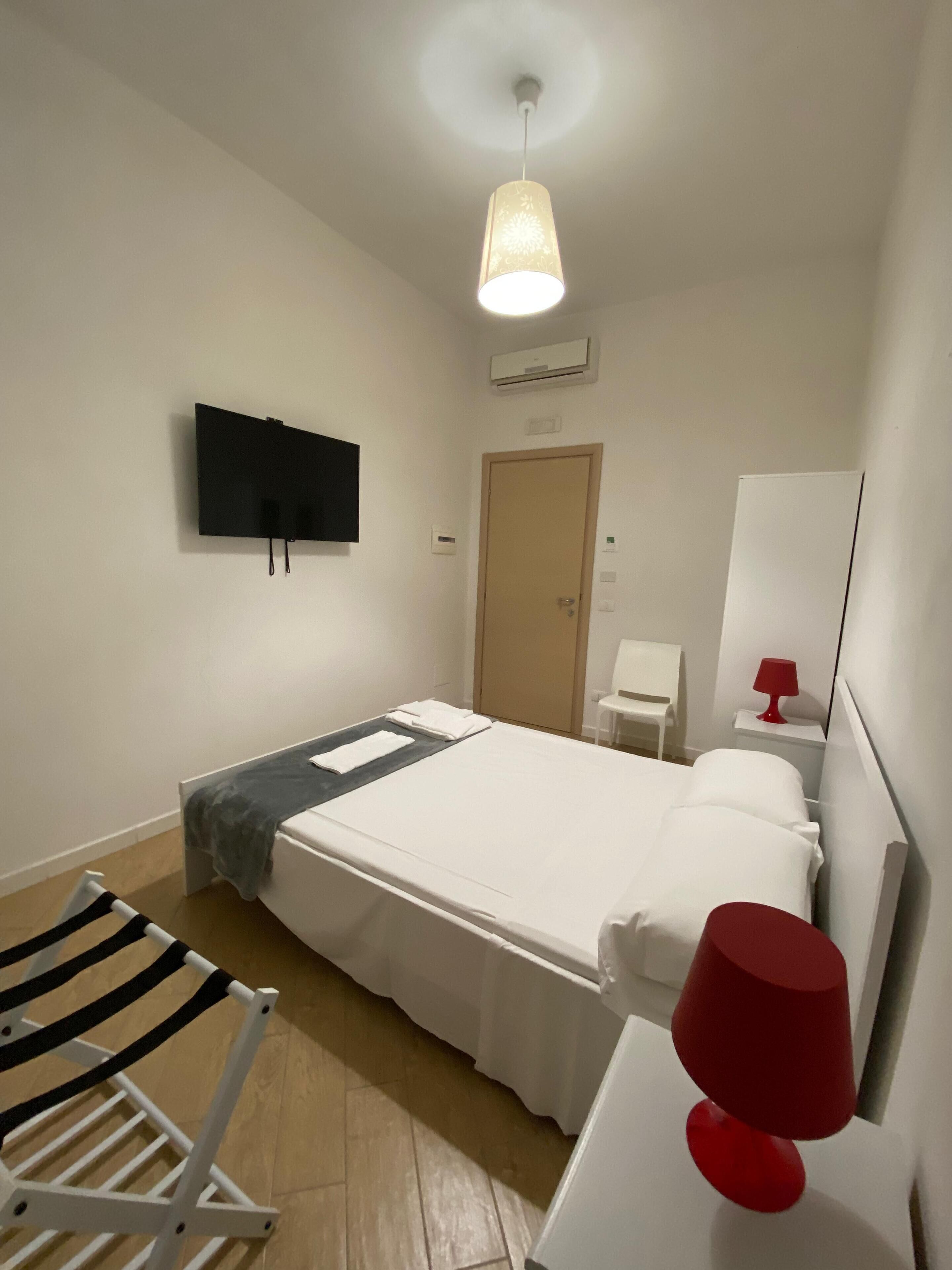 Une chambre d’hôtel avec un lit, un téléviseur fixé au mur, un bureau avec une lampe rouge et une chaise.
