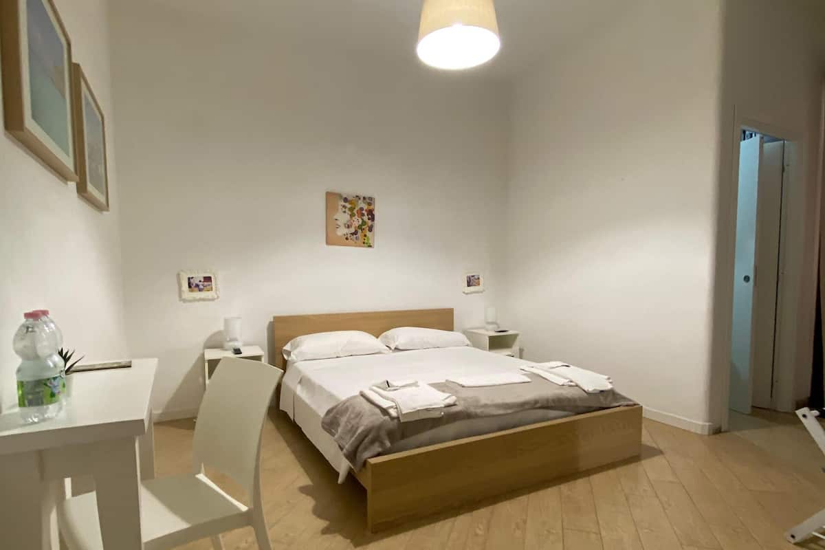 A bedroom with a wooden bed, white walls, a desk with a chair, and a hanging light fixture.