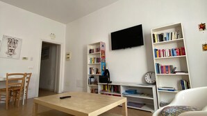A modern living room with a wooden coffee table, a white sofa with a colorful cushion, a bookshelf, a flat-screen TV, and a ceiling light fixture. - Manu's house B&B (Porto Empedocle)