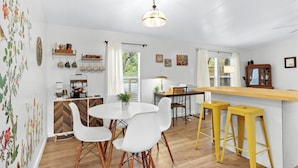 Een eetgedeelte met een witte tafel, stoelen en een gele barstoel. Er is een houten kast met een visgraatmotief, een hangplant en een raam met witte gordijnen.