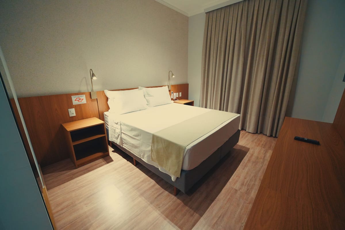 Une chambre d’hôtel avec un lit, des tables de chevet, une petite table, une lampe et un rideau.