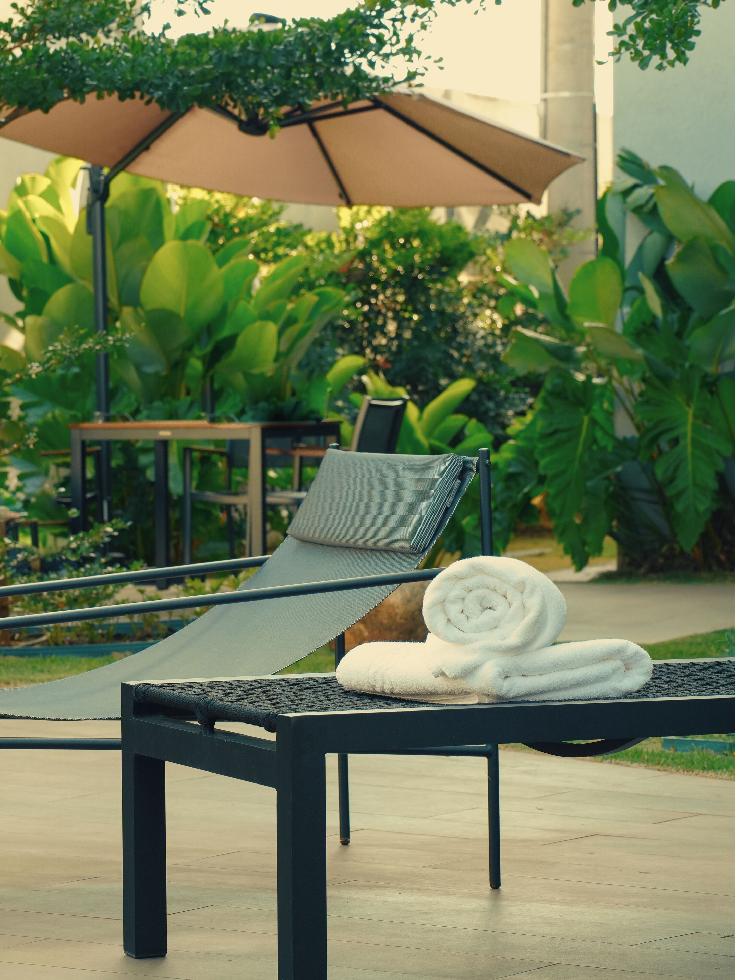 A lounge chair with a towel, a table, and an umbrella in a garden setting.