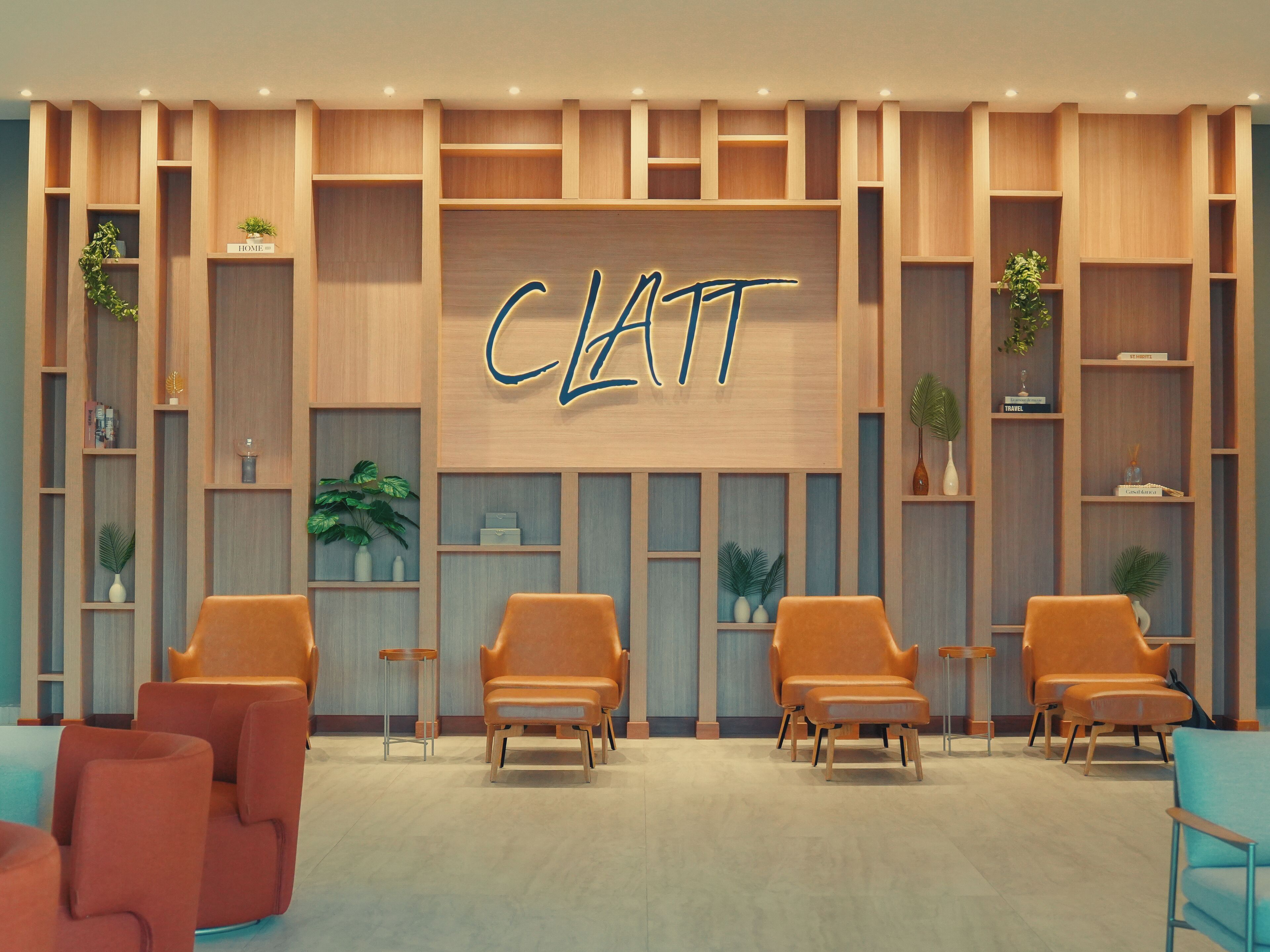 A modern interior with a central wall displaying the name 'CLATT' in illuminated letters, flanked by wooden shelving units with plants and decorative items, and several orange chairs arranged in the foreground.