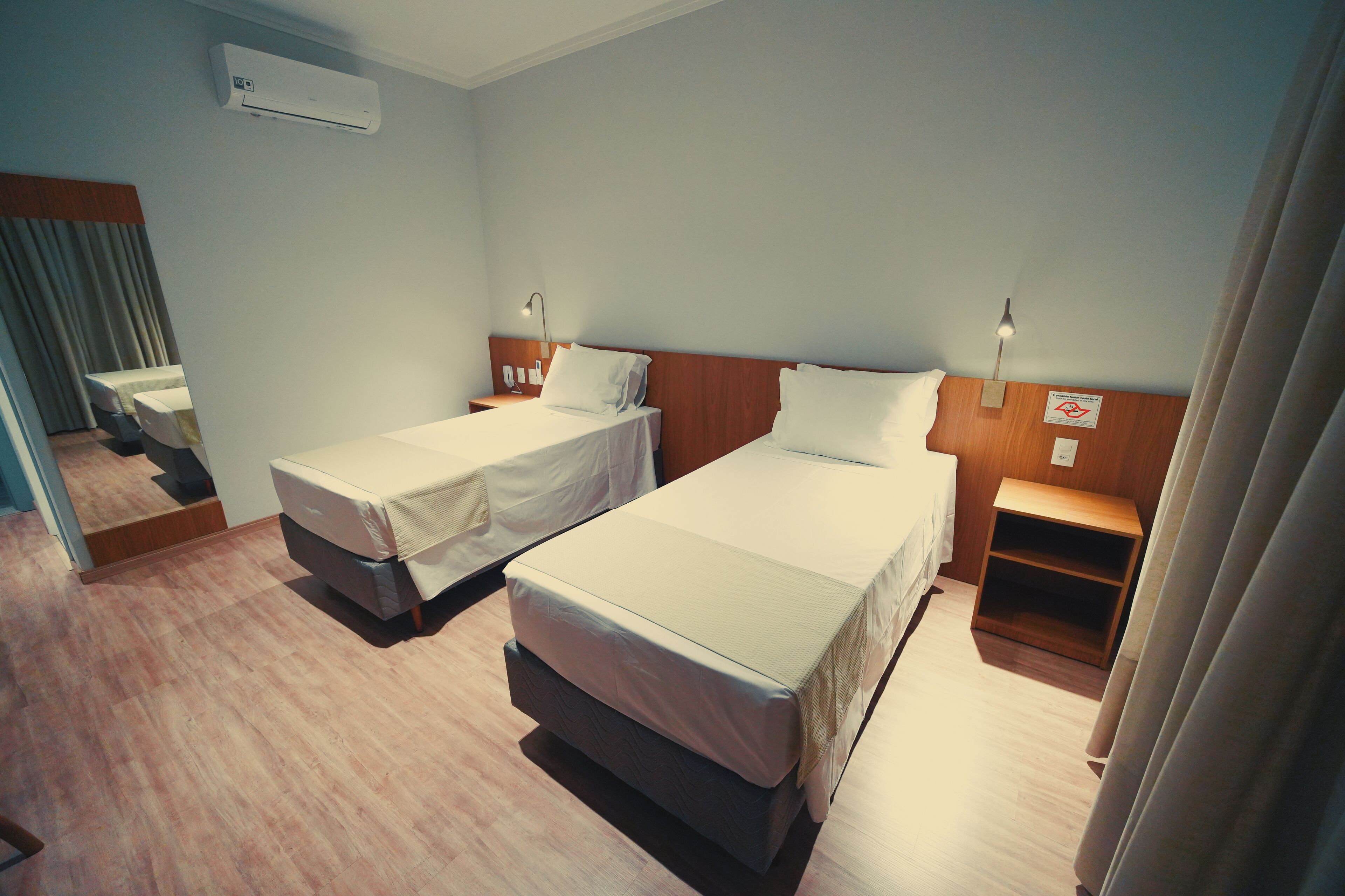 A hotel room with two single beds, a wooden nightstand, a wall-mounted air conditioner, and a small wooden shelf.