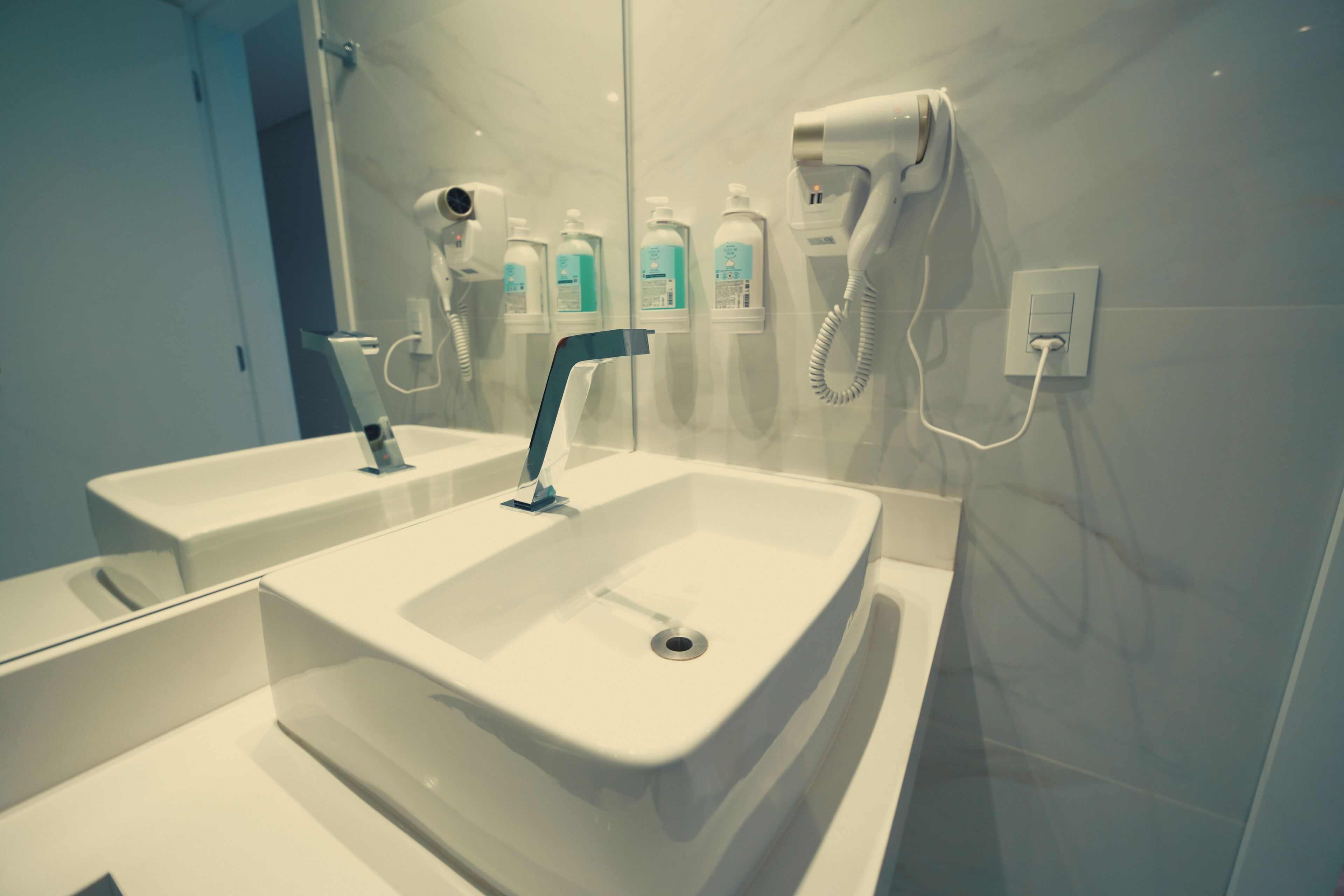 Une salle de bain moderne avec un grand miroir, deux lavabos, un sèche-cheveux et plusieurs flacons de produits de toilette accrochés au mur.