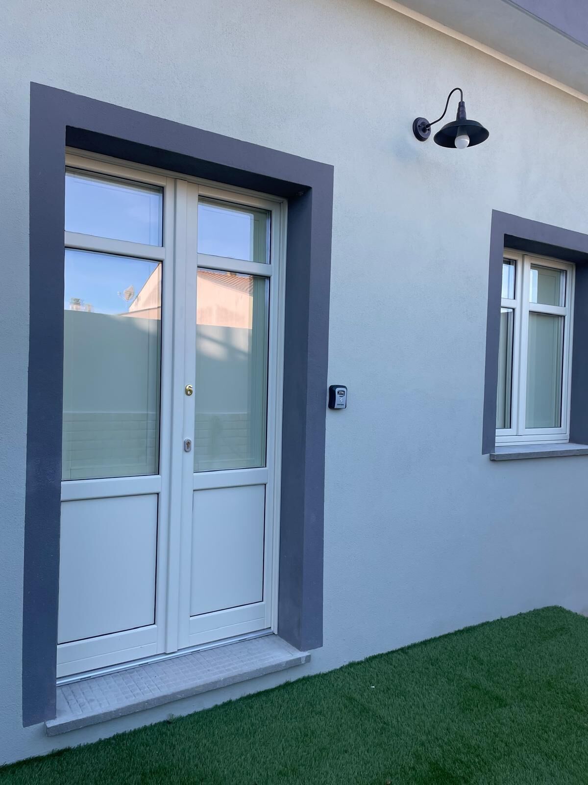 A modern building with a white door and window, a small outdoor area with artificial grass, and a wall-mounted light fixture.