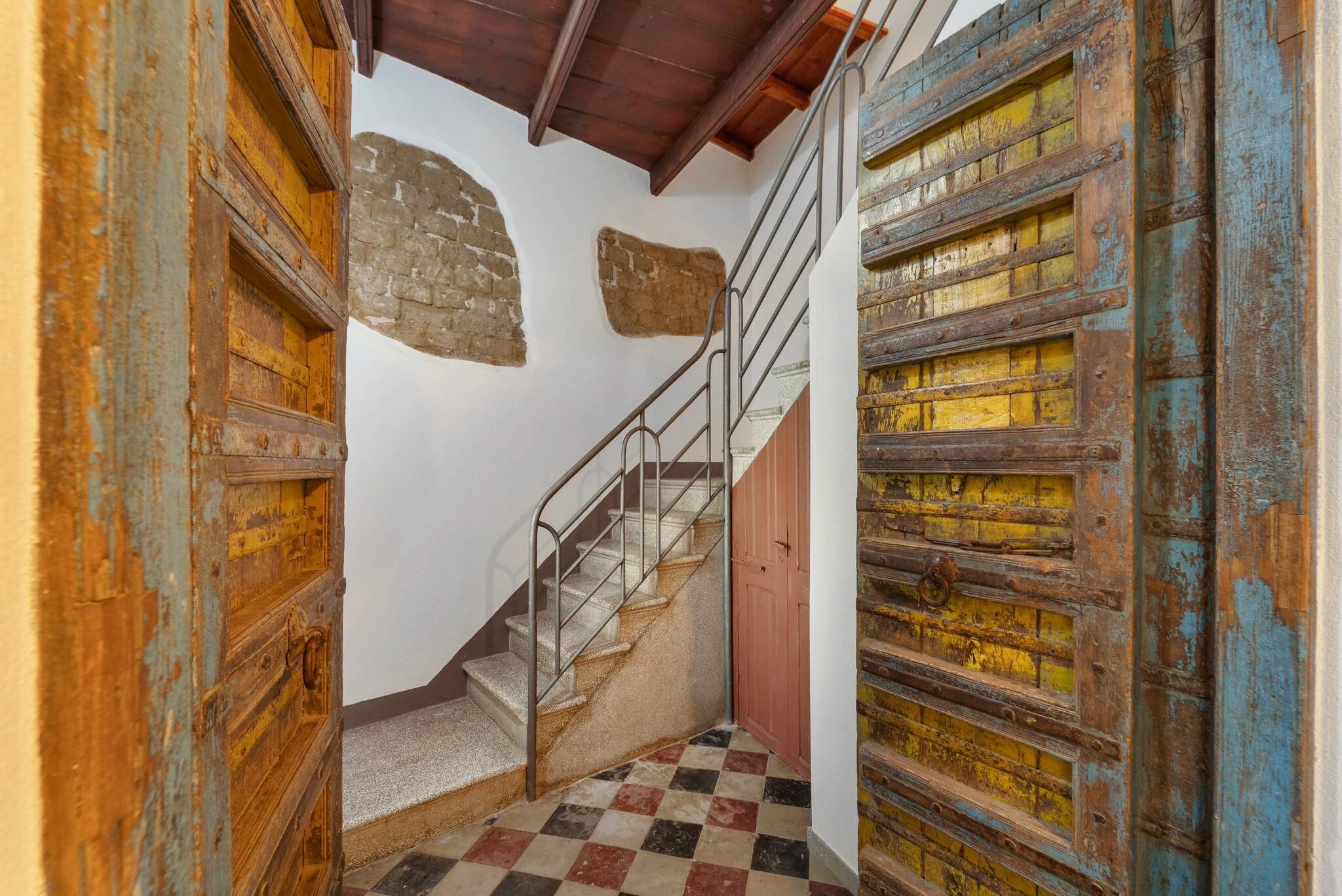 Un escalier avec des rampes en métal, un sol à carreaux et des portes en bois rustiques dont la peinture s’écaille.
