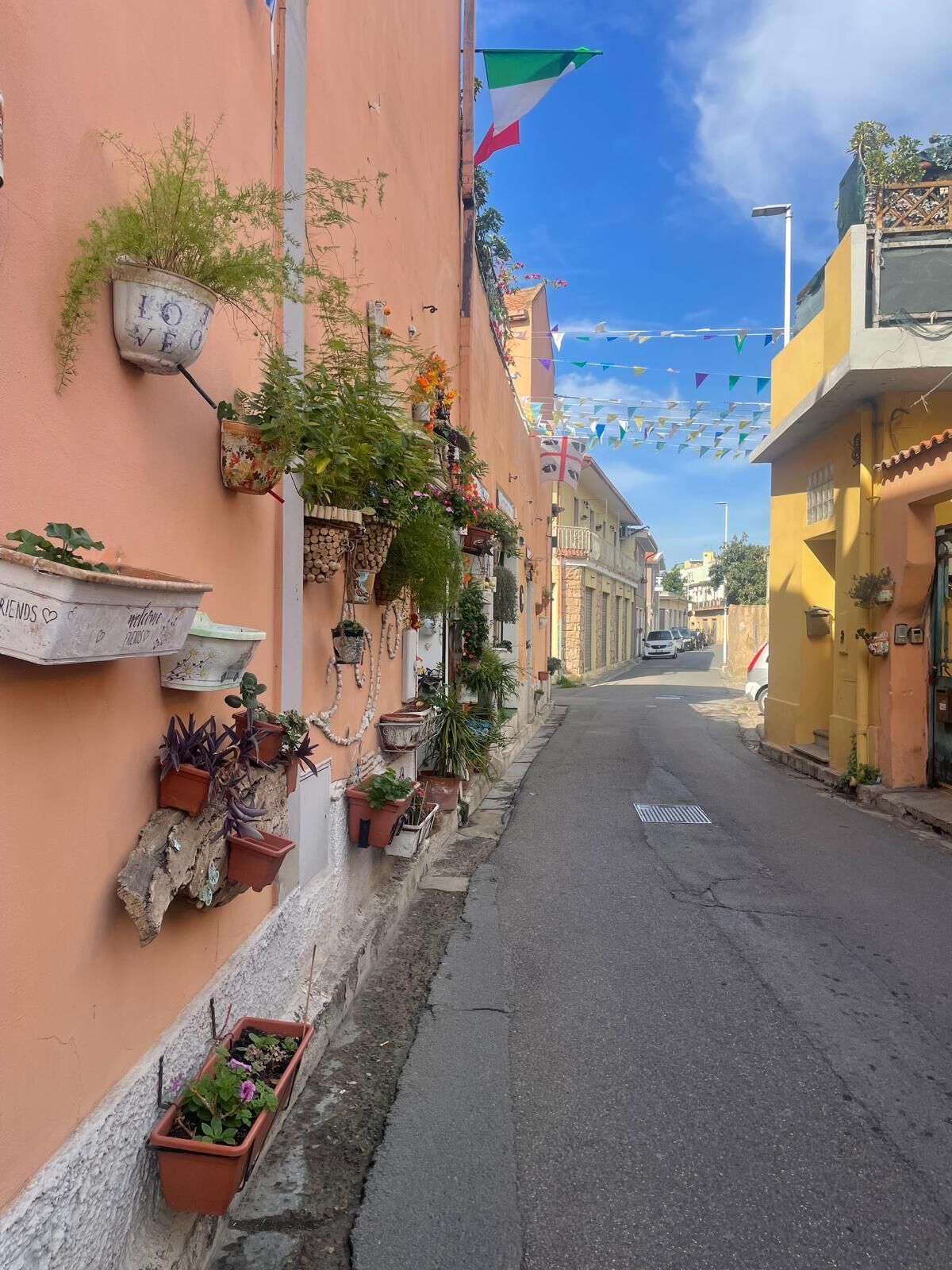 A narrow street lined with buildings, some with potted plants and flowers, and a clear blue sky above.