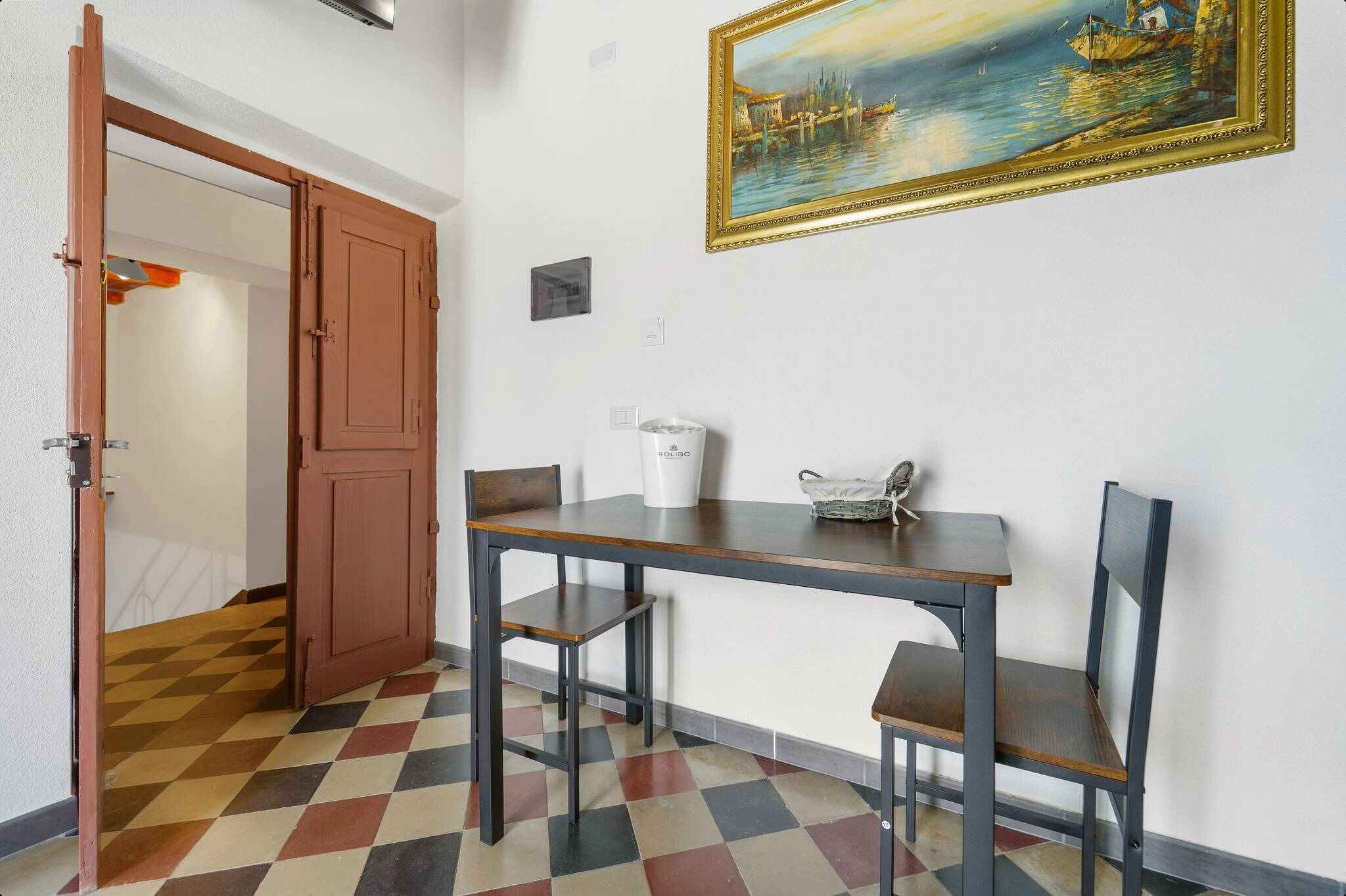 Een kamer met een geruit vloerkleed, een tafel met stoelen, een schilderij aan de muur en een deur naar een andere kamer.