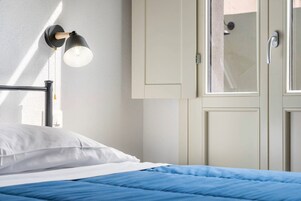 Una cama con ropa de cama blanca y azul, un aplique en la pared y una puerta con manija.