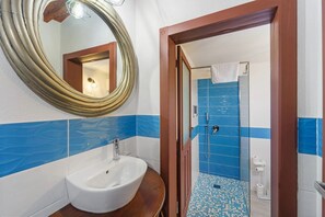 A bathroom with a white sink, a wooden vanity, a large mirror, and blue-tiled walls. - Luxar Suites & Apartments (Assemini)