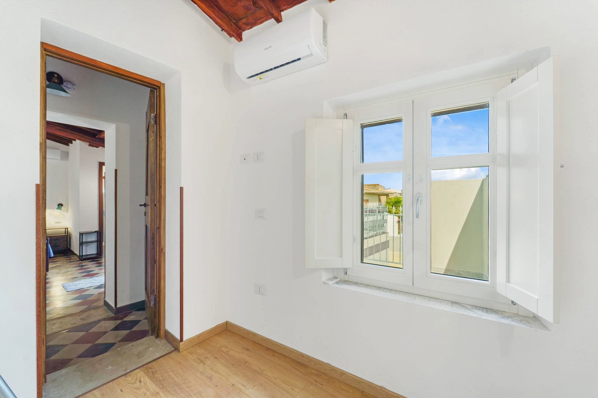 A room with a wooden floor, white walls, a window with white shutters, and an air conditioning unit.