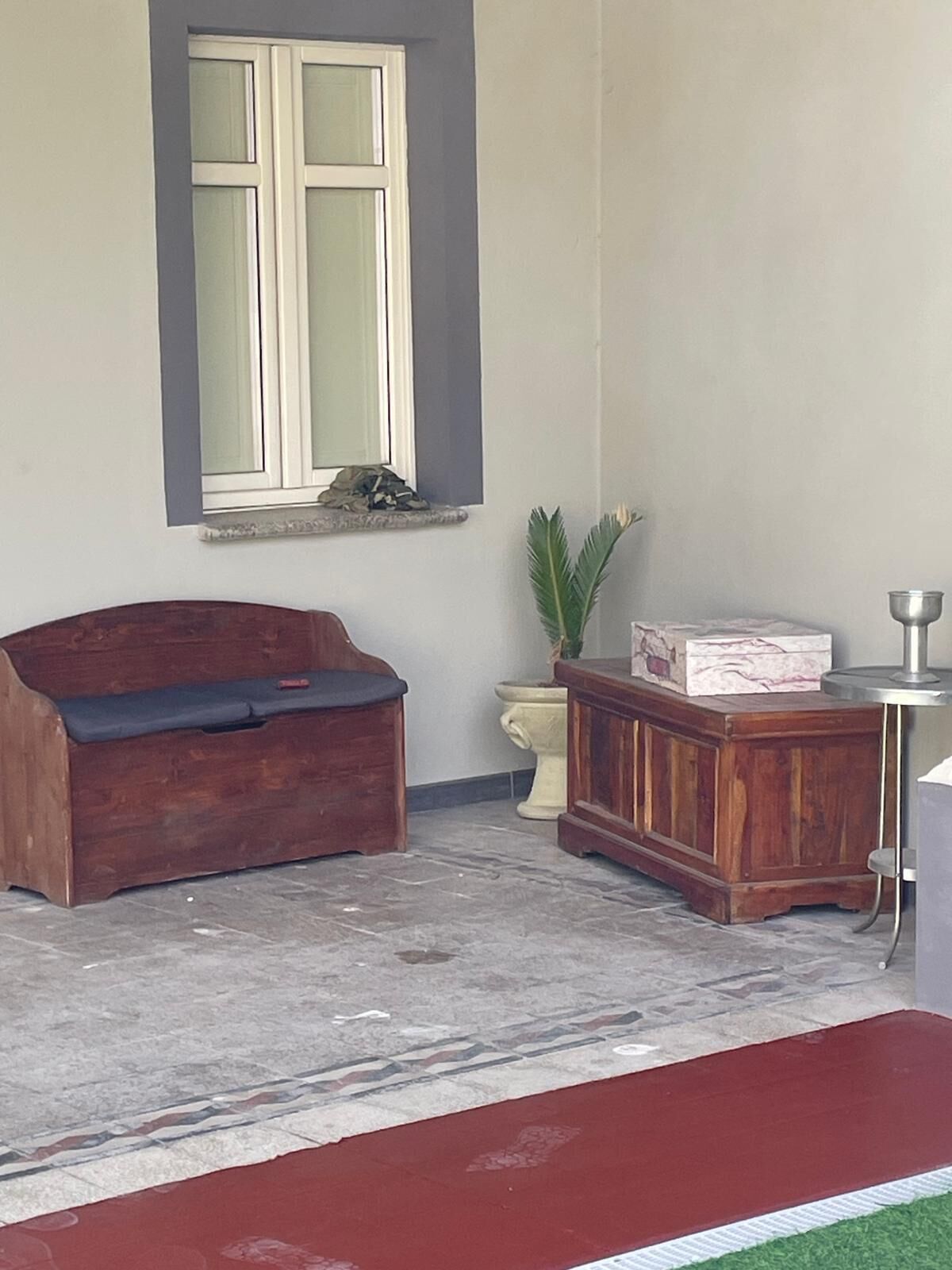 A room with a wooden bench, a small table with a metal cup, a potted plant, and a window with a curtain.
