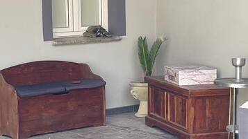 A room with a wooden bench, a small table with a metal cup, a potted plant, and a window with a curtain.