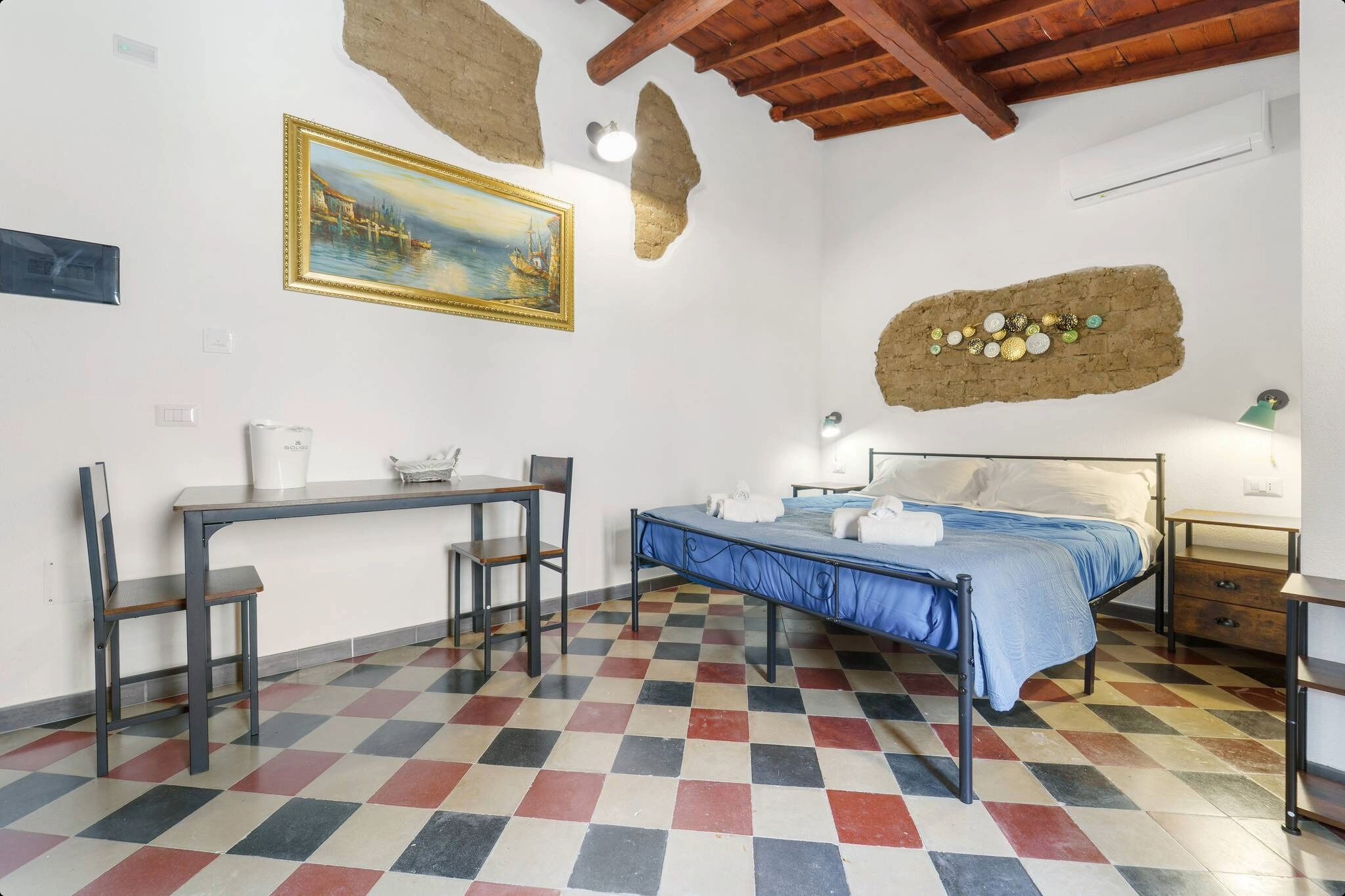 Una habitación con piso a cuadros, una cama con cubrecama azul, un escritorio con silla, un cuadro en la pared y un adorno rústico en la pared.