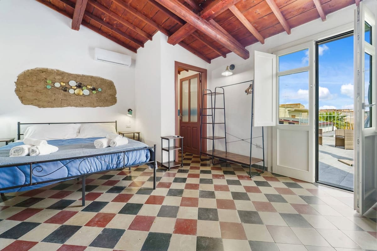 Ein Zimmer mit Schachbrettmuster auf dem Boden, einem Bett mit aufgerollten Handtüchern, einer Holzbalkendecke, einer Wanddekoration und einer zur Außenseite führenden Tür.