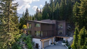 A modern two-story house with a garage, surrounded by dense forest and a stone retaining wall. - New luxury chalet with hot tub & media (Whistler)