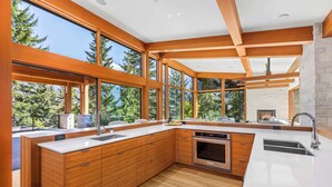 A modern kitchen with large windows, wooden cabinetry, and a stone fireplace. - New luxury chalet with hot tub & media (Whistler)