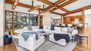 A spacious living area with a large sectional sofa, a coffee table, and a view of a forest through large windows. - New luxury chalet with hot tub & media (Whistler)